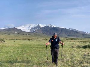 Person with walking poles standing in grasslands with a snow covered mountain range behind.
