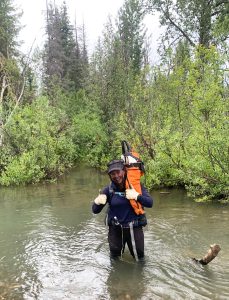 A person standing in a river knee deep, carrying a chainsaw wrapped in an orange bag.