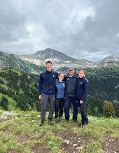 Four people smiling and standing on a mountain with forest and mountains behind.