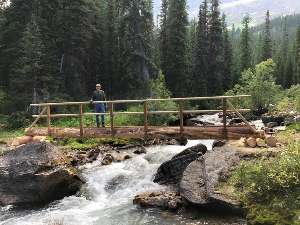 A volunteer stands on a newly-built wooden bridge over fast-moving water. Boulders dot the foreground, and trees and mountains are in the background. 