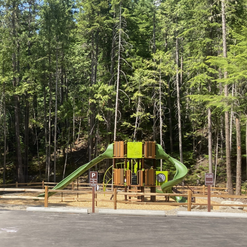 Photo of a wood and green plastic playground in front of a forest, with a new concrete parking lot in the foreground. 