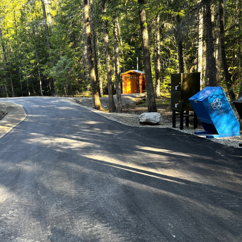 Photo of a newly paved road winding through a forested campground, new waste and recycling bins on the righthand side and a new outhouse in the distance.  