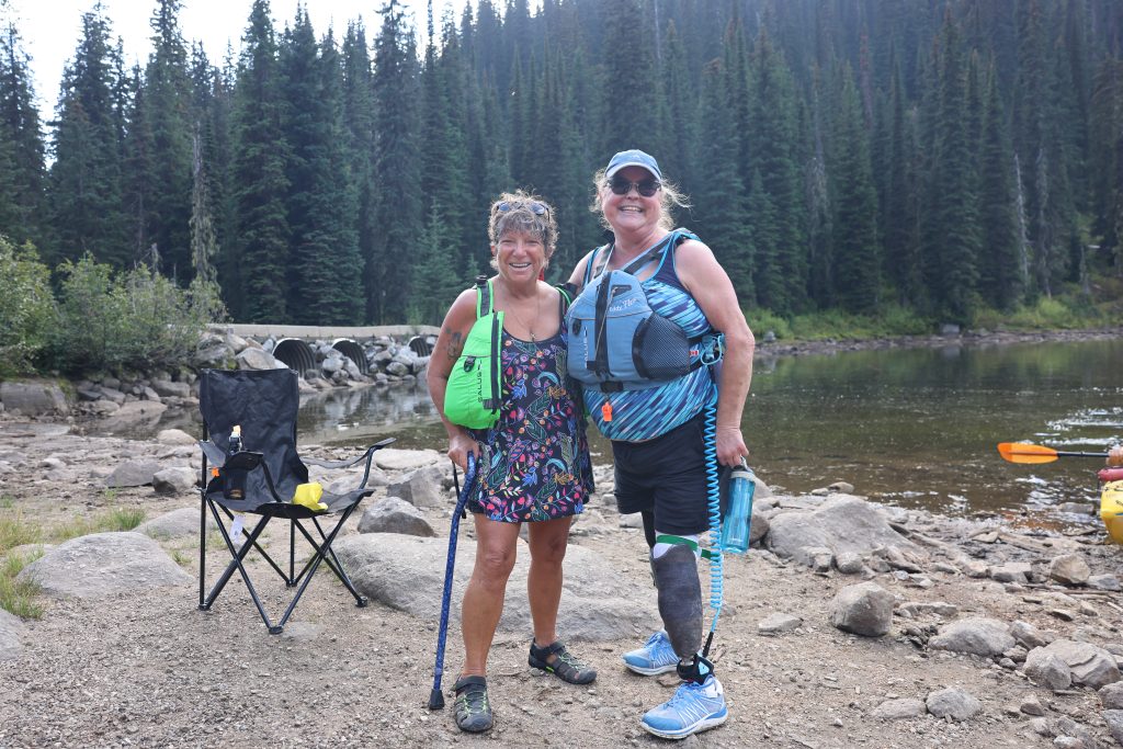 Two people standing on the lakeshore smiling at the camera. The person on the left is wearing a green life jacket and is holding a walking cane. The person on the left is wearing a blue life jacket and is wearing a prosthetic leg.