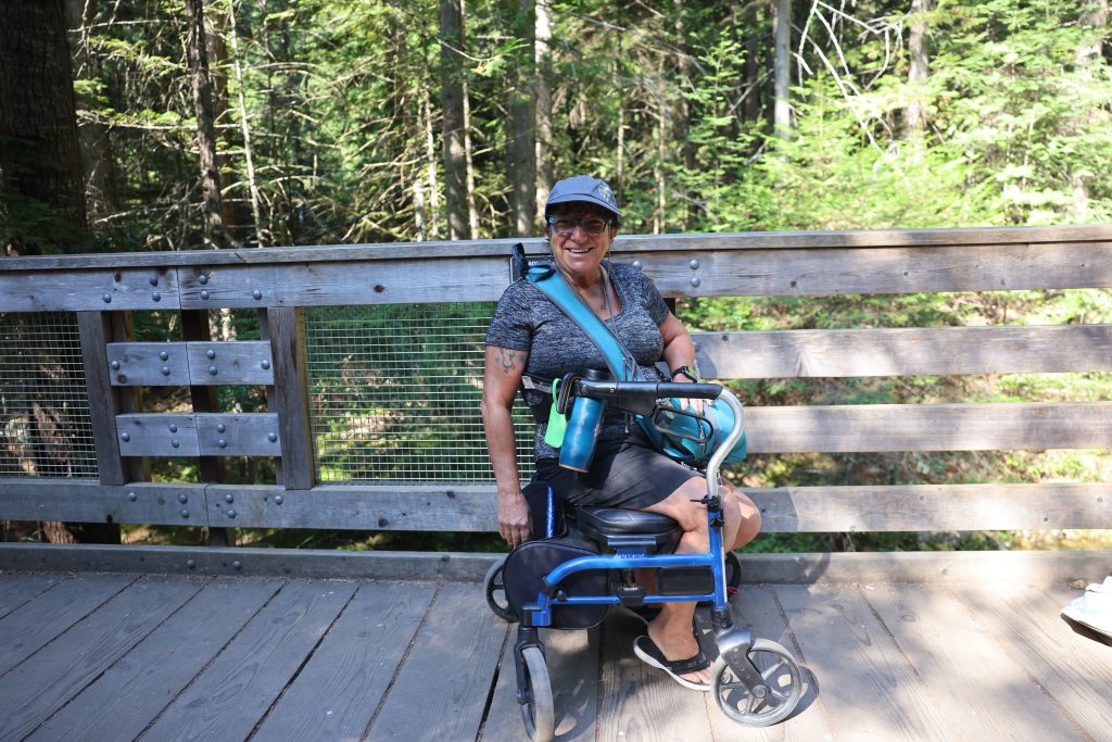 A person sitting on their walker smiling at the camera. They are sitting on a wooden bridge, and green trees are in the background.