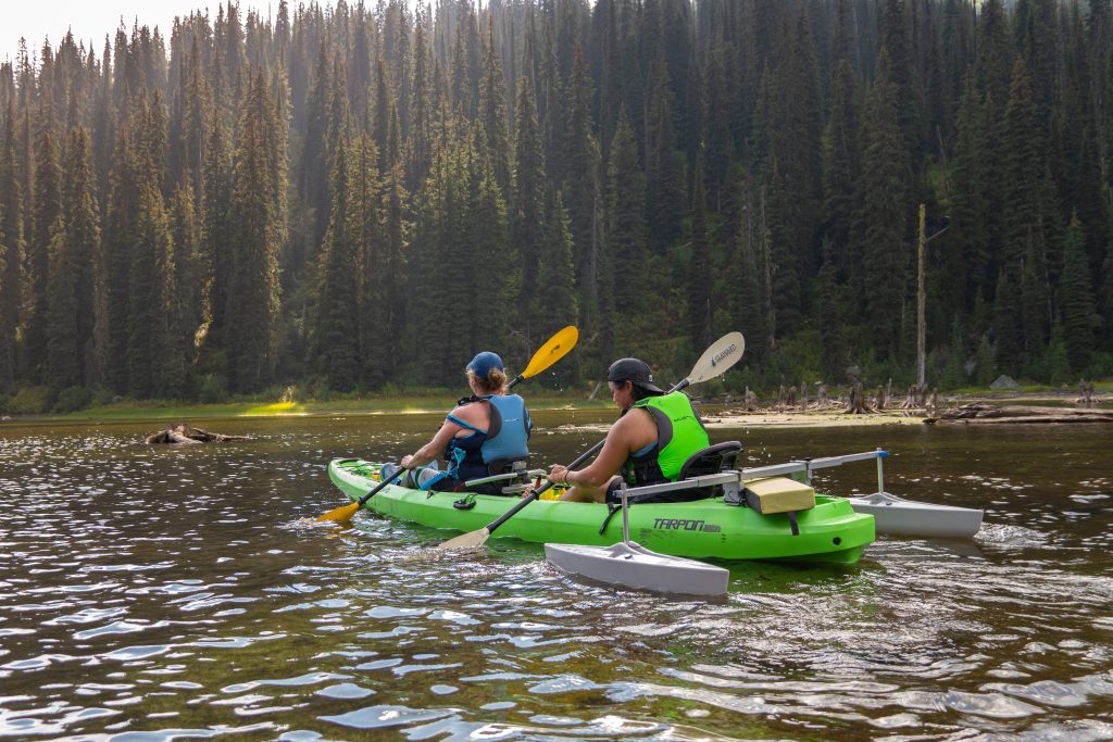 Two people paddling on a lake in a green kayak with outriggers. Green trees are in the background.