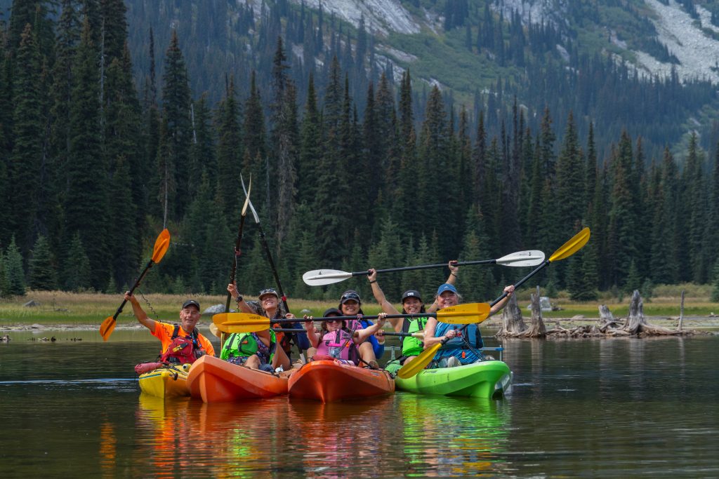  A group of seven people in four colourful kayaks lined up for a group photo on a lake with their paddles held in the air. Green trees and a towering mountain are in the background.