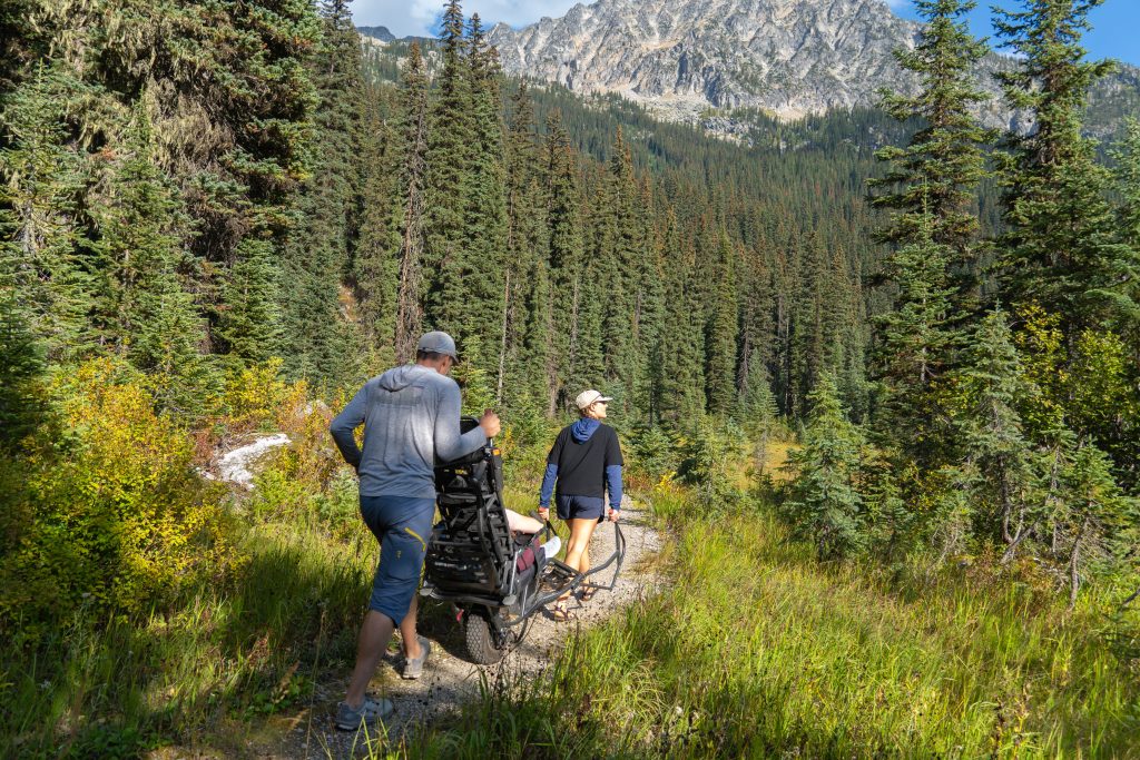 A person sitting in an adaptive hiking device which is being maneuvered by two people on a trail in the forest.