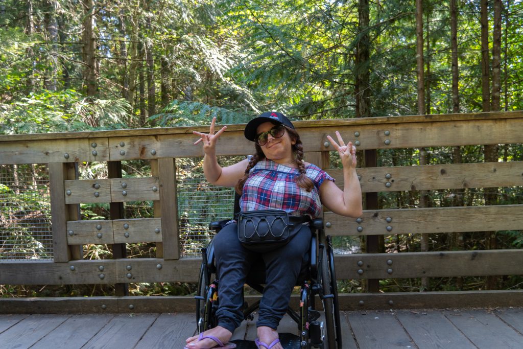 A manual wheelchair user posing for the camera on a wooden bridge. Green trees are in the background.