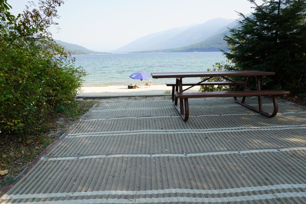 An accessible picnic table on an accessible mat at the edge of a beach. A view of a lake and mountains are in the background.